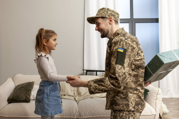 um soldado militar ucraniano masculino de uniforme voltou para casa e abraça sua filha, um pai patriótico dá um presente para a criança e comemora seu aniversário - Foto, Imagem