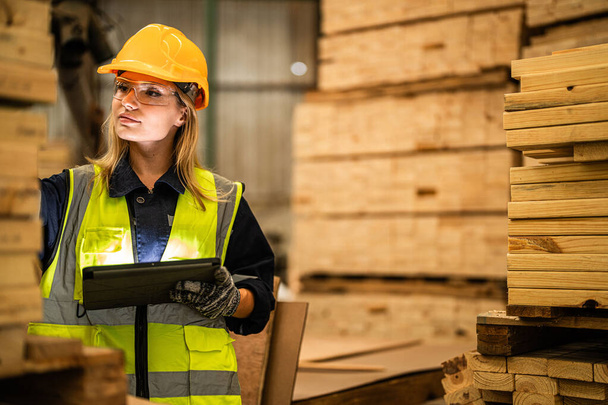 γυναίκα ξυλουργός εργαζόμενος φορώντας στολή ασφαλείας και σκληρό καπέλο εργασίας και τον έλεγχο της ποιότητας των ξύλινων προϊόντων κατά την κατασκευή του εργαστηρίου. άνδρες και γυναίκες εργαζόμενοι ξύλο στη σκοτεινή βιομηχανία αποθηκών. - Φωτογραφία, εικόνα