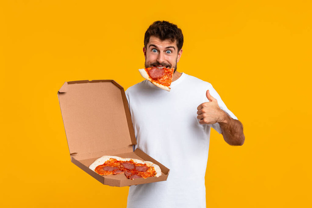Happy Guy met plak pizza in de mond Gesturing duimen omhoog op gele Studio achtergrond, goedkeuring Pizzeria levering en smaak, glimlachen om de camera. Grote Junk Food Concept, Cheat Meal - Foto, afbeelding
