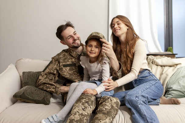 young soldier of the Ukrainian army in a camouflage uniform returned home to his family, a military dad hugs his little daughter and wife at home, a happy military family sits at home on the sofa and - Photo, Image