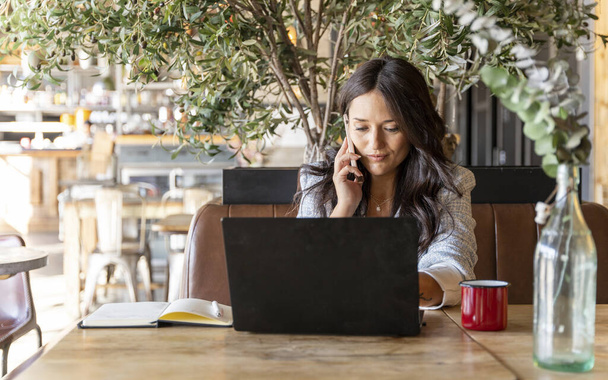 喫茶店でラップトップコンピュータでオンラインで働いている間,女性が電話で話す. テクノロジーとビジネスコンセプト. - 写真・画像