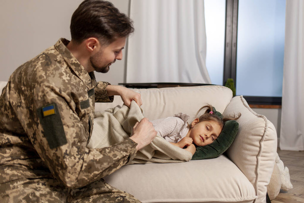 mladý voják ukrajinské armády v maskovací uniformě se vrátil domů ke svému dítěti, vojenský otec přikryje a políbí svou dcerku na pohovku doma, koncept vojenské Ukrajiny - Fotografie, Obrázek