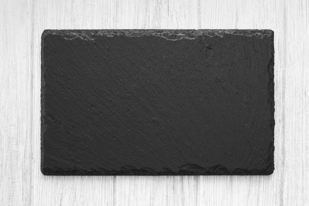 自然なスレートの石の空の板が付いている白い木のテーブルの背景. 食糧供給のための装飾的な黒い版. - 写真・画像