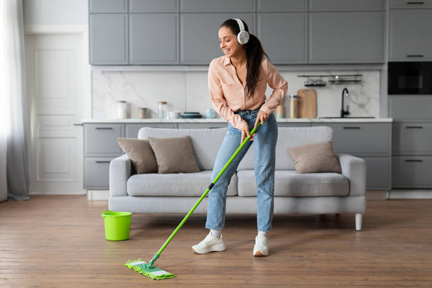 Woman in pastel shirt and jeans happily mopping her wooden floor while listening to music with white headphones, accompanied by a green bucket - Photo, Image