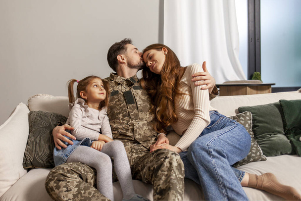 jonge soldaat van het Oekraïense leger in een camouflage uniform terug naar huis naar zijn familie, een militaire vader knuffelt zijn kleine dochter en vrouw thuis, een gelukkig militair gezin zit thuis op de bank en - Foto, afbeelding