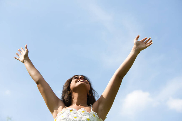 Красивая испанская брюнетка с длинными вьющимися волосами поднимает руки к голубому небу. Женщина счастлива и благодарит Бога и жизнь за каждый день. Понятие счастья и благодарности - Фото, изображение