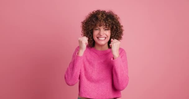 Statische Echtzeit-Aufnahme einer aufgeregten jungen Frau mit lockigem Haar in Make-up und Rundhalsausschnitt, die lässig in die Kamera schaut, während sie im Stehen klatschend in die Hände klatscht und Erfolg auf rosa Hintergrund feiert - Filmmaterial, Video