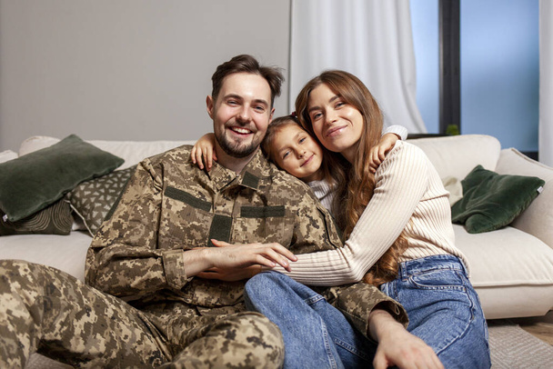 Молодой солдат украинской армии в камуфляжной форме вернулся домой к семье, военный папа обнимает дочку и жену дома, счастливая военная семья сидит дома на диване и - Фото, изображение