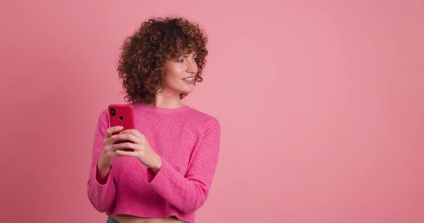 Mujer joven sonriente en traje rosa con el pelo rizado que navega por el teléfono inteligente mientras apunta hacia el fondo rosa - Metraje, vídeo