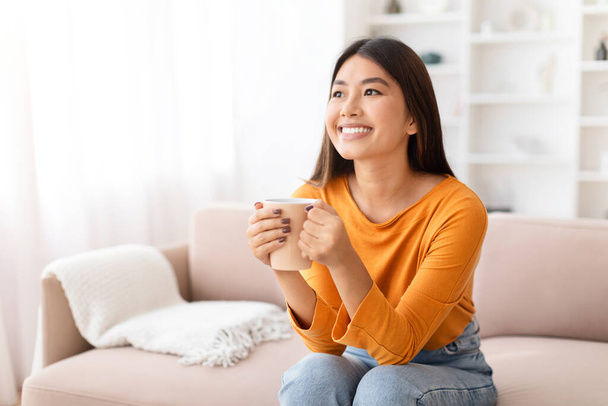 Портрет щасливої привабливої молодої азіатки в повсякденному одязі, що сидить на дивані, тримає кухоль, п'є каву вдома, дивлячись на простір копіювання та посміхаючись. Домашнє дозвілля, відпочинок - Фото, зображення