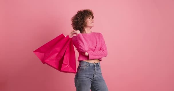 ピンクの背景に立っている間,ピンクの紙のショッピングバッグを保持するカーリーヘアでカジュアルな服で若い女性を笑う - 映像、動画