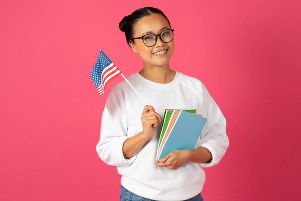 Lächelnde asiatische Studentin, stolz mit amerikanischer Flagge und bunten Notizbüchern, fröhliche junge Koreanerin, die für internationale Bildung und kulturellen Austausch steht, steht auf rosa Hintergrund - Foto, Bild