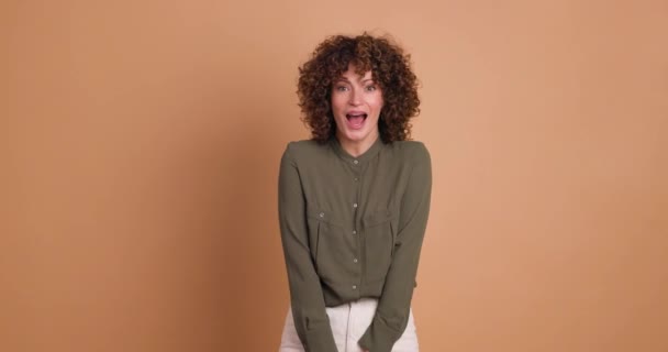 Statische Echtzeit-Aufnahme einer erstaunten jungen Frau mit gelocktem Haar, Make-up und offenem Mund, die in die Kamera schaut, während sie mit gefalteten Händen steht und Haare auf beigem Hintergrund berührt - Filmmaterial, Video