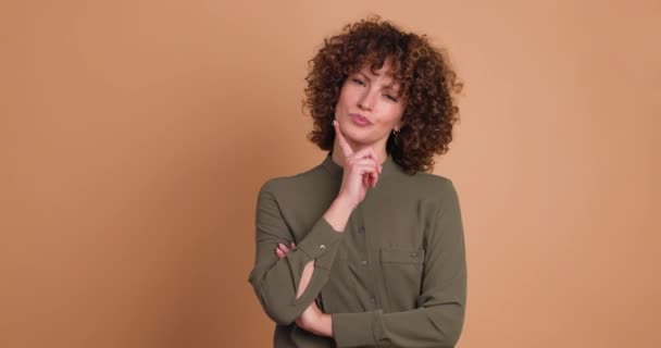 Fotografía estática en tiempo real de una joven y pensativa hembra de pelo rizado en maquillaje y una mirada casual a la cámara mientras está de pie con la mano cruzada y tocando la barbilla con el dedo sobre un fondo beige - Metraje, vídeo