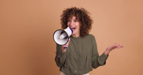 Mujer joven alegre en ropa casual con el pelo rizado gritando en megáfono, mientras que de pie sobre fondo beige - Imágenes, Vídeo