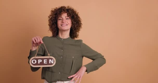 Curly haired woman with open sign in hand, with a smile on a beige background. - Footage, Video