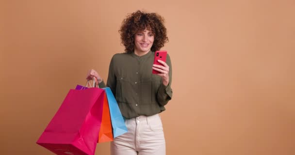 Удивлённая молодая женщина в повседневной одежде с вьющимися волосами, держащая разноцветные бумажные пакеты и используя смартфон, стоя на бежевом фоне - Кадры, видео
