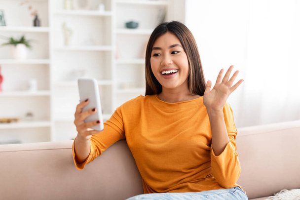 Positive pretty young asian woman sitting at couch at home, waving and smiling at smartphone screen, have video call with family or friends, copy space, telecommunication concept - Photo, Image