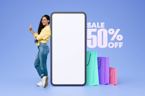 Shopping Discount Angebot. Lächelnde arabische junge Käuferin posiert neben leerem Handybildschirm mit Einkaufstaschen, Werbung zu 50 Prozent vor blauem Studiohintergrund. Collage - Foto, Bild