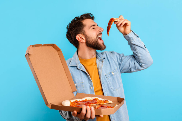 ピザ・ローバー。 ジャンクフードを重視するおかしい男, ブルースタジオの背景にボックスでポーズピザのスライスを食べる, 不健康な栄養習慣を紹介. ガイ エンジイ チート ミール コンセプト - 写真・画像