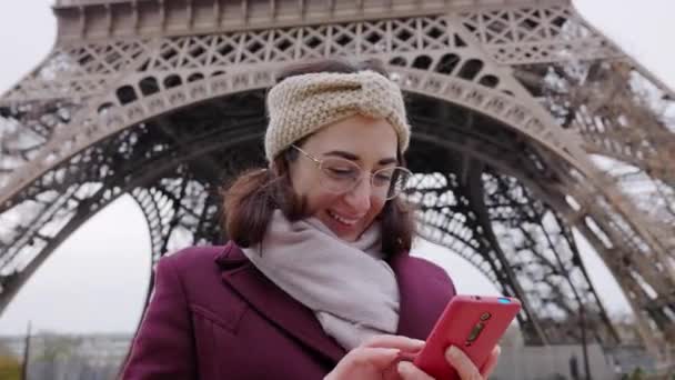 Νεαρή ευτυχισμένη γυναίκα που χρησιμοποιεί κινητό τηλέφωνο μπροστά από τον Πύργο του Άιφελ στο Παρίσι, Γαλλία. Έννοια ταξιδιών και τεχνολογίας. - Πλάνα, βίντεο
