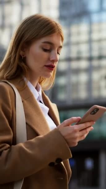 Pionowy widok eleganckiej kobiety przedsiebiorczyni czytajacej wiadomosci finansowe przez telefon podczas przechodzenia przez przejscie poprzeczne w megalopolis. Pewna siebie bizneswoman przechodząca przez ulicę i SMS-y przez telefon - Materiał filmowy, wideo