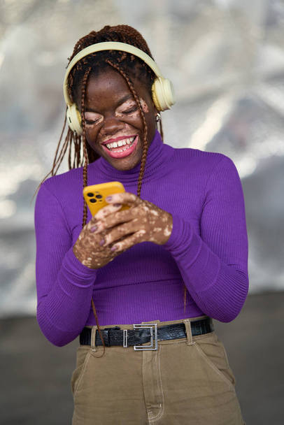 Сияющая женщина с витилиго наслаждается мелодиями на наушниках, взаимодействуя со своим желтым телефоном - Фото, изображение
