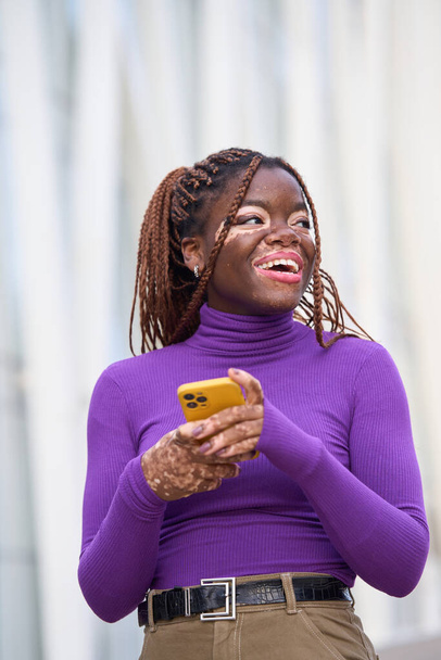 Una mujer alegre con vitiligo mira hacia arriba mientras escribe en su teléfono inteligente amarillo, vestido con un suéter púrpura ceñido - Foto, imagen