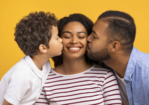 Adorable son and loving husband giving kisses on cheeks of smiling black mother, showing love and affection, greeting their mom and wife with birthday or womens day, standing on yellow background - Photo, Image