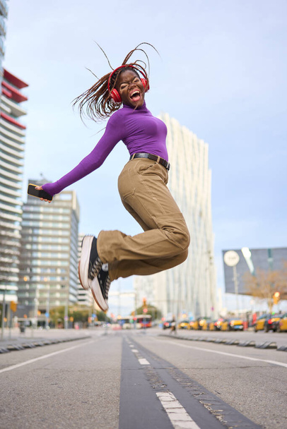 An ecstatic young woman with vitiligo and braids leaps high, listening to music in the heart of the city - Photo, Image