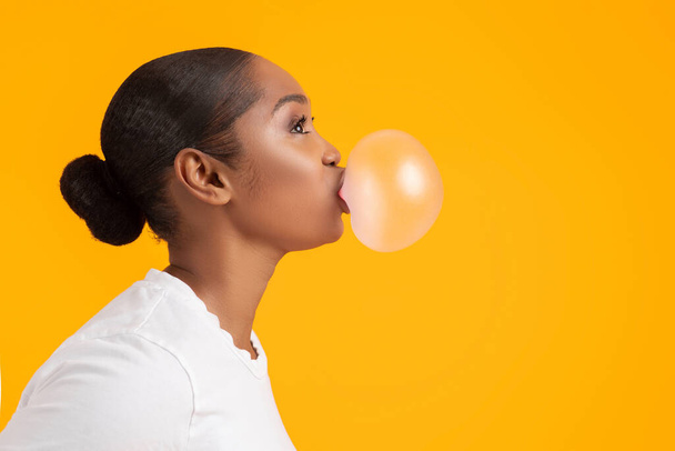 Вид сбоку портрет черной молодой женщины, надувающей пузырь, смотрящей в сторону на копировальное пространство на желтом фоне студии. Крутая афроамериканка тысячелетия жующая жвачку, сладкоежка - Фото, изображение