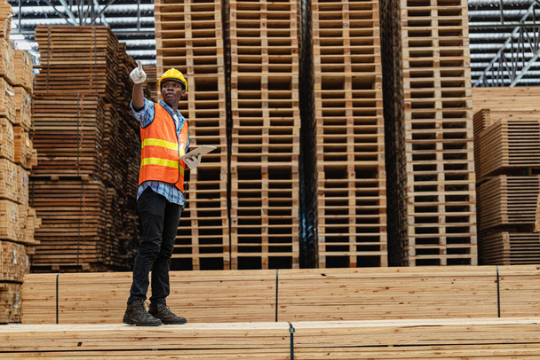 Αφρικανοί εργαζόμενοι άνθρωπος μηχανική περπάτημα και επιθεώρηση με την εργασία σουίτα φόρεμα και γάντι χειρός σε αποθήκη ξύλου. Έννοια της έξυπνης βιομηχανίας εργαζόμενος που λειτουργεί. Εργοστάσια ξύλου παράγουν ουρανίσκο ξύλου. - Φωτογραφία, εικόνα