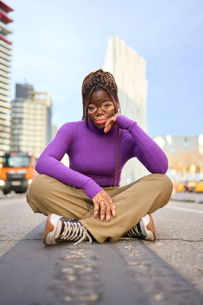 Μια στοχαστική μαύρη γυναίκα με λεύκη κάθεται σε ένα δρόμο της πόλης, το βλέμμα της στοχαστικό μέσα στο αστικό τοπίο. - Φωτογραφία, εικόνα