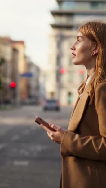 Вертикальный взгляд позитивной бизнес-леди, читающей сообщение мобильного телефона во время перехода дороги на солнечной улице, общающейся в Интернете с друзьями во время перерыва на работу - Кадры, видео