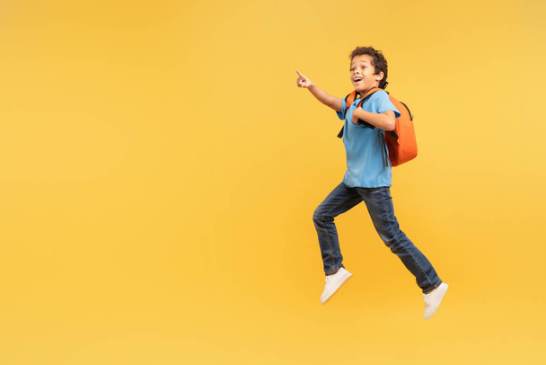Lively boy with curly hair in mid-stride pointing forward at free space with excitement, wearing blue shirt and carrying backpack on bright yellow background, full of energy and adventure - Photo, Image