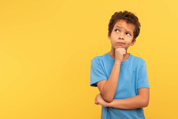 Thoughtful young boy with curly hair looking aside at copy space, touching chin in contemplation, wearing blue shirt on yellow background, embodying curiosity and deep thought - Photo, Image