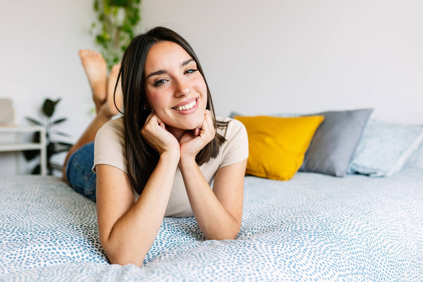 Lächelndes Porträt einer jungen, fröhlichen Frau, die auf dem Bett liegt und in die Kamera blickt. Entspannte Frau zeigt zahmes Lächeln, das auf dem Schlafzimmer ruht. - Foto, Bild