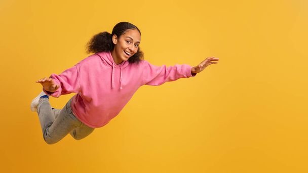 Ευτυχισμένο μαύρο κοριτσάκι με ροζ κουκούλα απλώνει τα χέρια και πετάει στον αέρα, απολαμβάνοντας την ελευθερία και τη διασκέδαση στα όνειρά της πάνω από το κίτρινο φόντο του στούντιο. Πλήρες πλάνο, πανόραμα με κενό χώρο - Φωτογραφία, εικόνα
