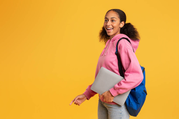 Возбужденная африканская студентка-студентка с ноутбуком и рюкзаком на фоне жёлтого студийного фона выражает радость по поводу начала учебного семестра. Отличное образовательное предложение. Копирование пространства - Фото, изображение