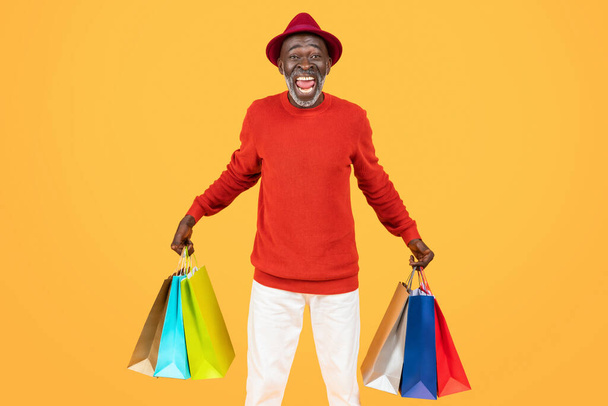 Alegre hombre afroamericano senior con vibrante atuendo rojo levanta triunfalmente coloridas bolsas de compras, capturando el espíritu de una experiencia de compra exitosa y agradable - Foto, imagen