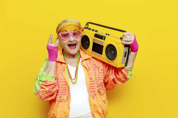 oude grappige oma in een jeugd kostuum danst met een tape recorder en luistert naar muziek op geel geïsoleerde achtergrond, oudere vrouw gepensioneerde in stijlvolle outfit zingt en toont rock gebaar op feest - Foto, afbeelding
