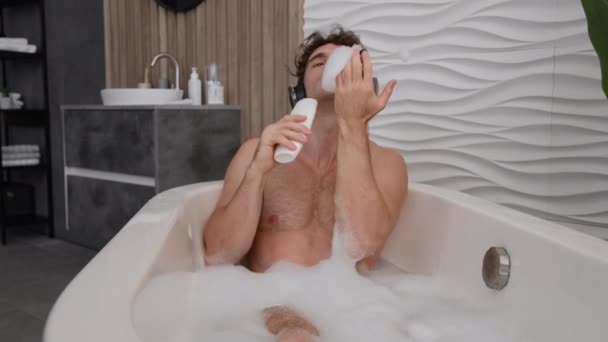 Despreocupado positivo tonto divertido caucásico hombre desnudo chico solo relajarse en el baño en agua de baño caliente disfrutar de casa descansando canto en gel de ducha como micrófono escuchando música en los auriculares - Imágenes, Vídeo