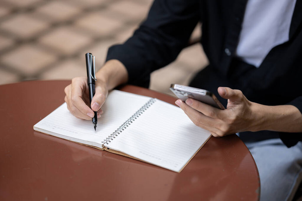 Imagem de perto de um homem usando seu smartphone e tomando notas em seu livro em uma mesa em um café. Conceito de estilo de vida - Foto, Imagem