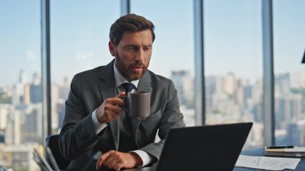 Зрілий лідер викликає ноутбук, який п'є каву вранці. Надійний чоловік розмовляє комп'ютером онлайн, працюючи в офісі на міському пейзажі. Корпоративний бородатий менеджер у костюмі, що просіює напій на відеоколекції - Кадри, відео