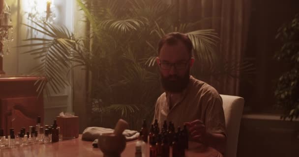 V reálném čase vousatý samec chemik v brýlích sedí u stolu a voní voňavý olej při výběru aroma pro nové parfémy v tmavé místnosti s matným světlem - Záběry, video