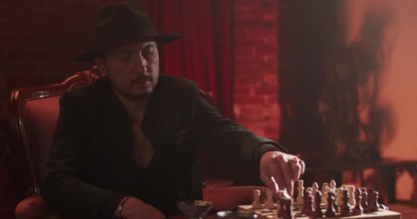 Άνθρωπος με αυτοπεποίθηση με καπέλο και κοστούμι που παίζει σκάκι με ανώνυμο φίλο σε σκοτεινό δωμάτιο στο σπίτι - Πλάνα, βίντεο