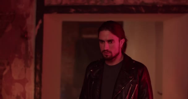 Продуманий дорослий бородатий чоловік у шкіряній куртці стоїть у темній кімнаті вдома під час освітлення - Кадри, відео