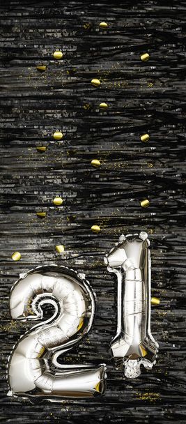 Globo de papel de plata número 21 sobre fondo de oropel negro decorado con confeti dorado. Tarjeta de felicitación de cumpleaños, inscripción veintiún. Evento de aniversario. Banner vertical. espacio de copia. - Foto, Imagen