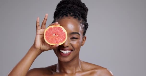 Grapefruit, schoonheid en gezicht van een vrolijke vrouw in de studio voor vitamine C voordelen, detox of gloed op grijze achtergrond. Portret, Afrikaans model of lach met citrusvruchten voor huidverzorging, duurzaamheid of dieet. - Video