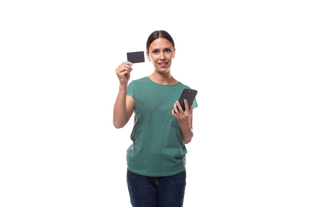 μια νεαρή γυναίκα με μαύρα μαλλιά και μια λεπτή φιγούρα ντυμένη με πράσινο μπλουζάκι διαφημίζει μια εκπτωτική κάρτα με μια μακέτα που κρατά ένα τηλέφωνο στο χέρι της. - Φωτογραφία, εικόνα